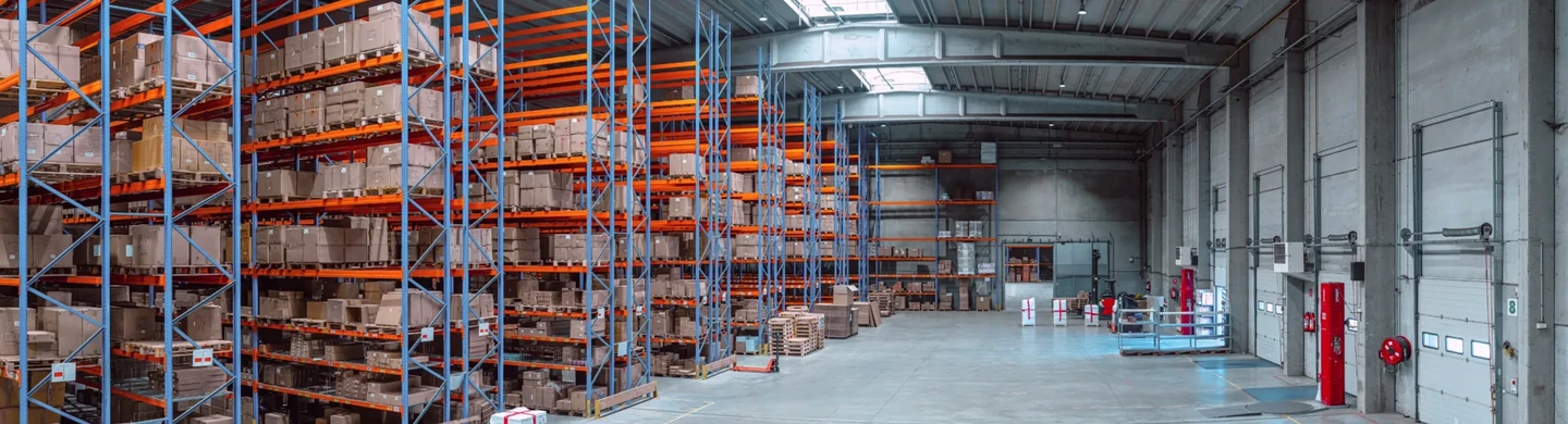 A warehouse with tall blue and orange pallet racks stacked with boxes and loading dock doors along the right wall