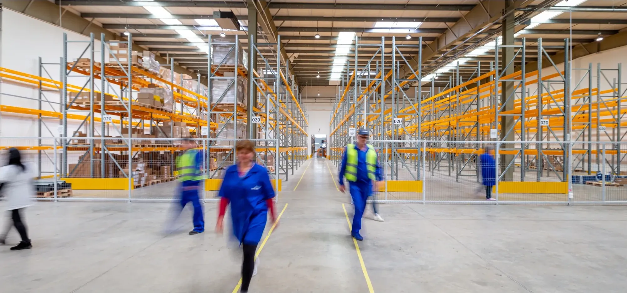 Workers in blue uniforms and high-visibility vests walking down a warehouse aisle between orange pallet racks