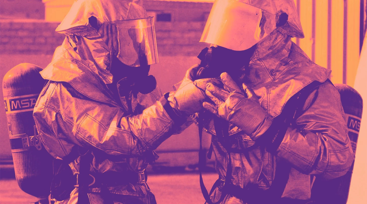 Two people in hazmat suits and breathing gear, one adjusting the other's mask